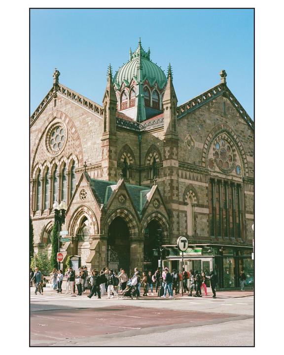 A large stone building with a green dome stands at a busy intersection where many people are crossing the street.