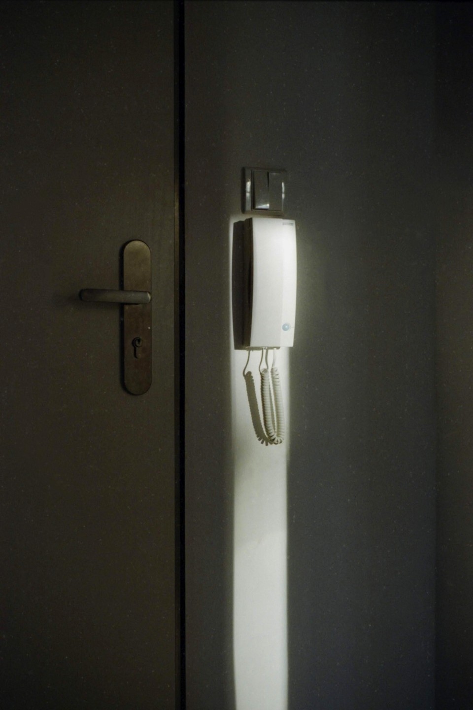 A white intercom is mounted on a wall next to a closed door, illuminated by a vertical beam of light.
