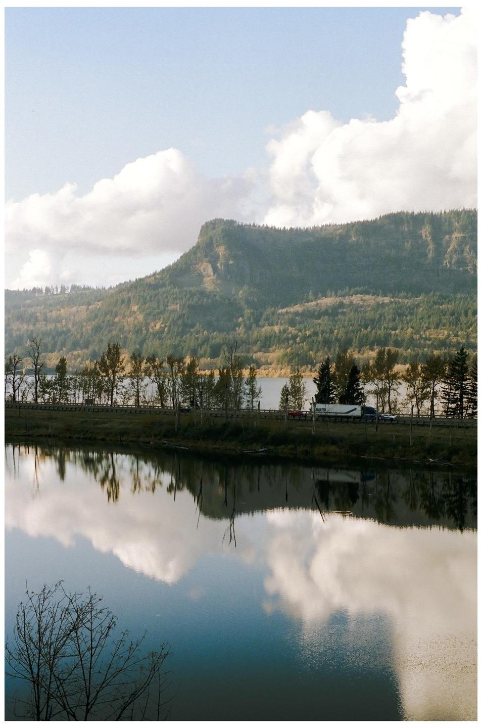 A body of water reflects the sky and a tree-covered mountain with a road and vehicles at its base.