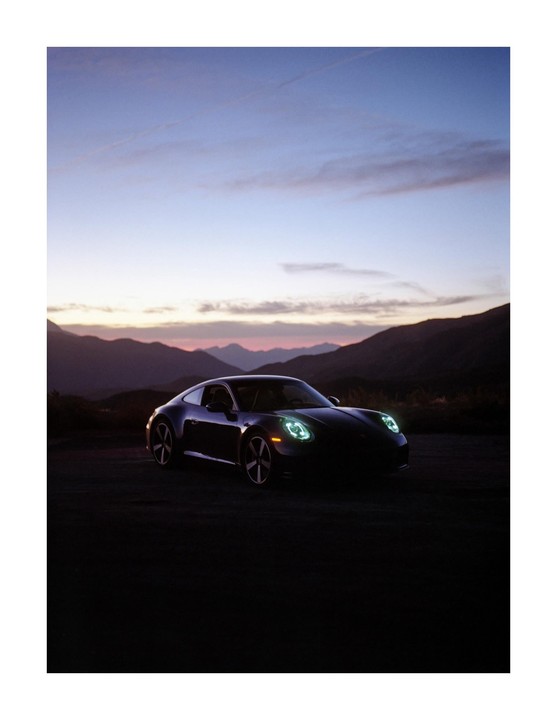 A dark car with its headlights on is parked in front of mountains at dusk.