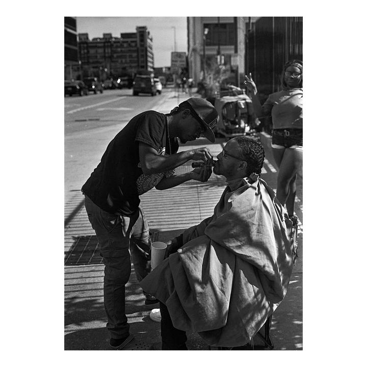 A man trims another man's beard on a city street while a woman stands in the background.