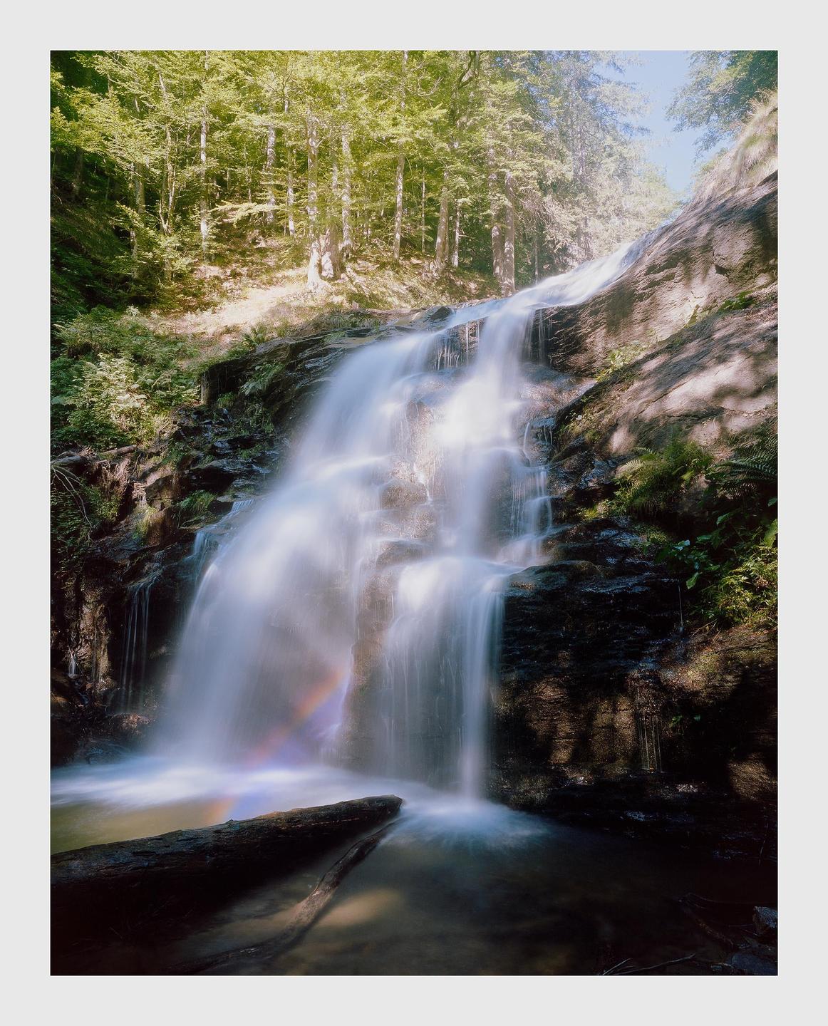 A long exposure photograph captures a waterfall cascading over rocks in a forest, with a rainbow visible in the mist and a fallen log in the foreground.