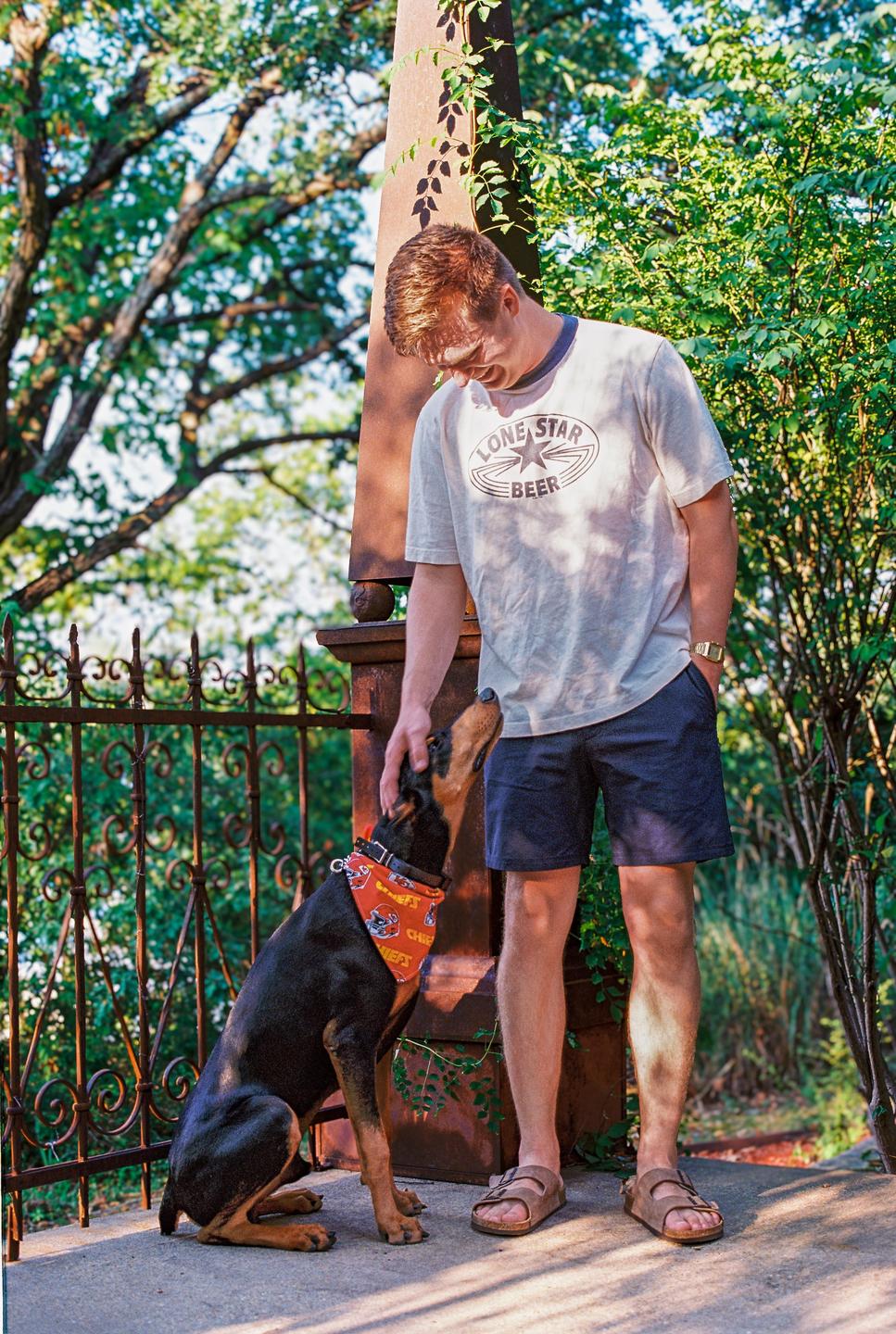A man wearing a t-shirt and shorts pets a doberman dog wearing a bandana next to a metal fence and a stone pillar.