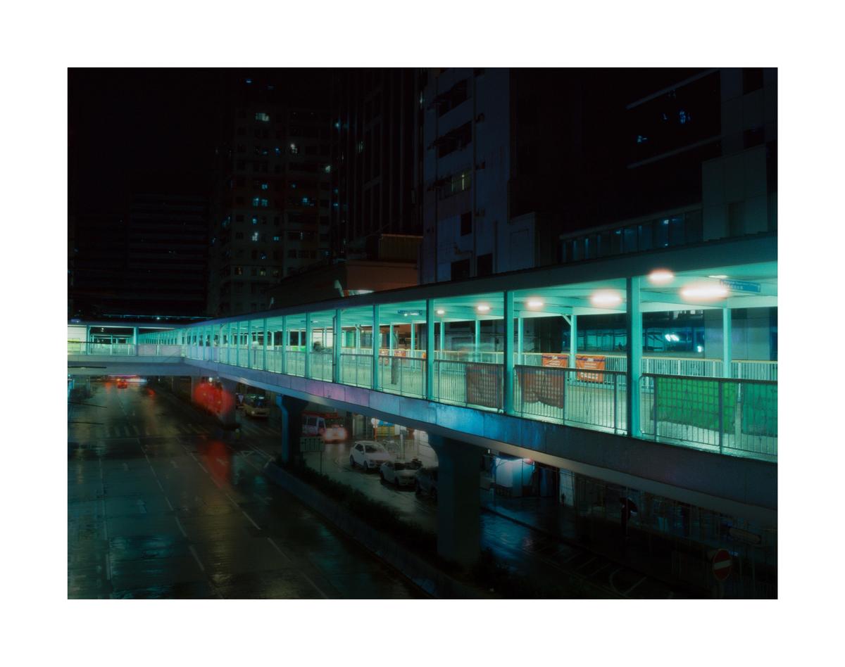 A long, illuminated pedestrian bridge stretches over a wet street with cars and buildings in the background at night.