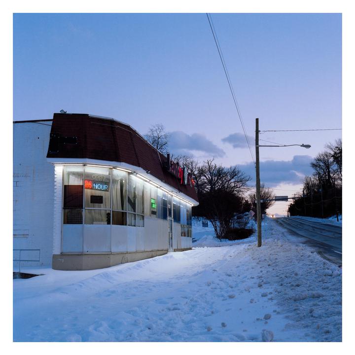 A brightly lit building with an "open 24 hour" sign stands next to a snow-covered road at dusk.