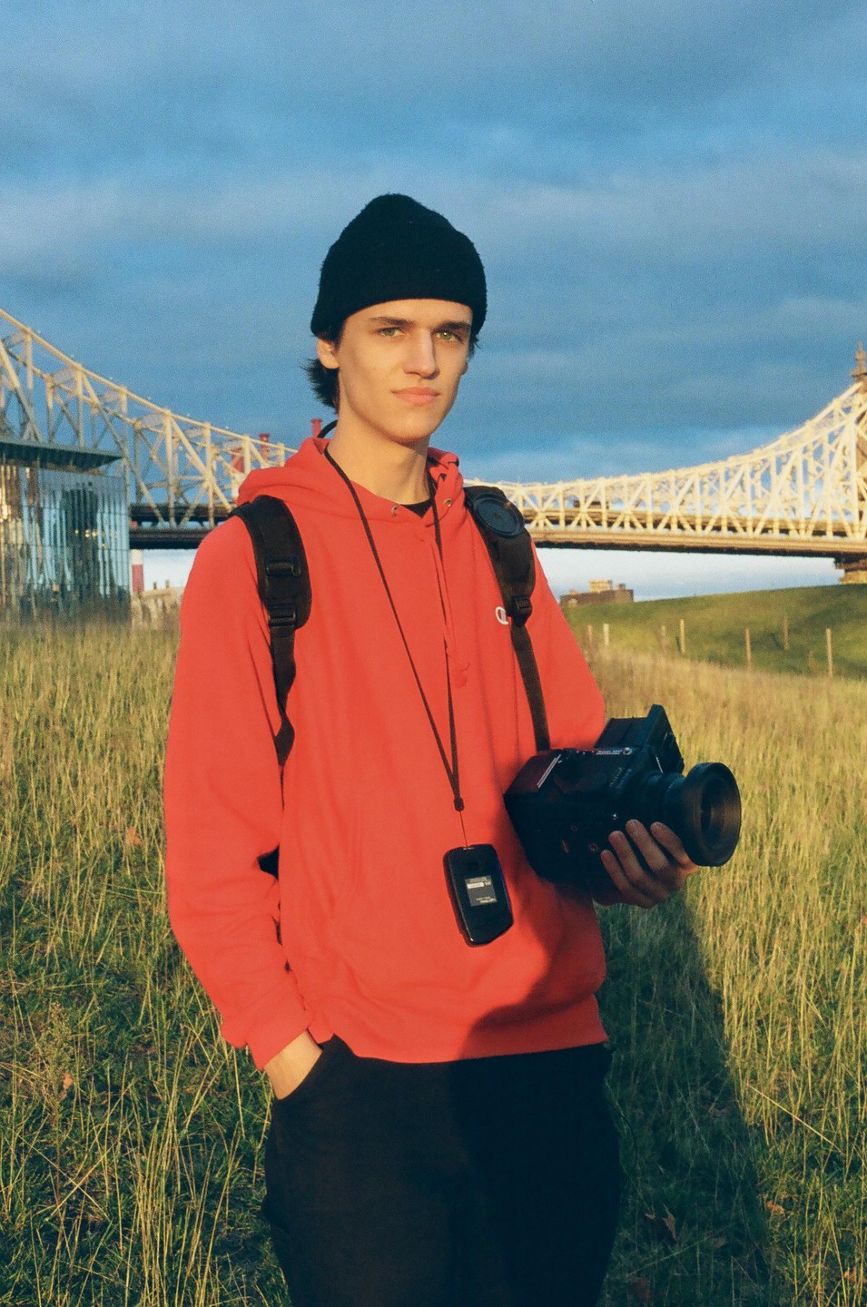 A young man in a red hoodie and black beanie holds a video camera in a grassy field with a bridge in the background.