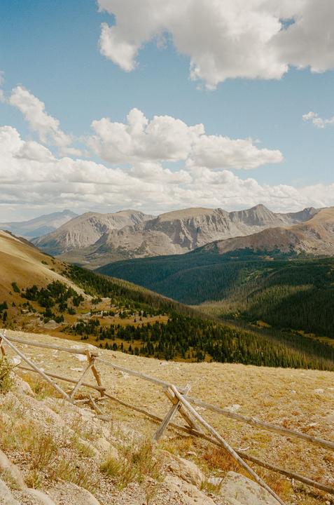 A wooden fence runs along a mountain slope with a valley and more mountains in the distance under a cloudy sky.
