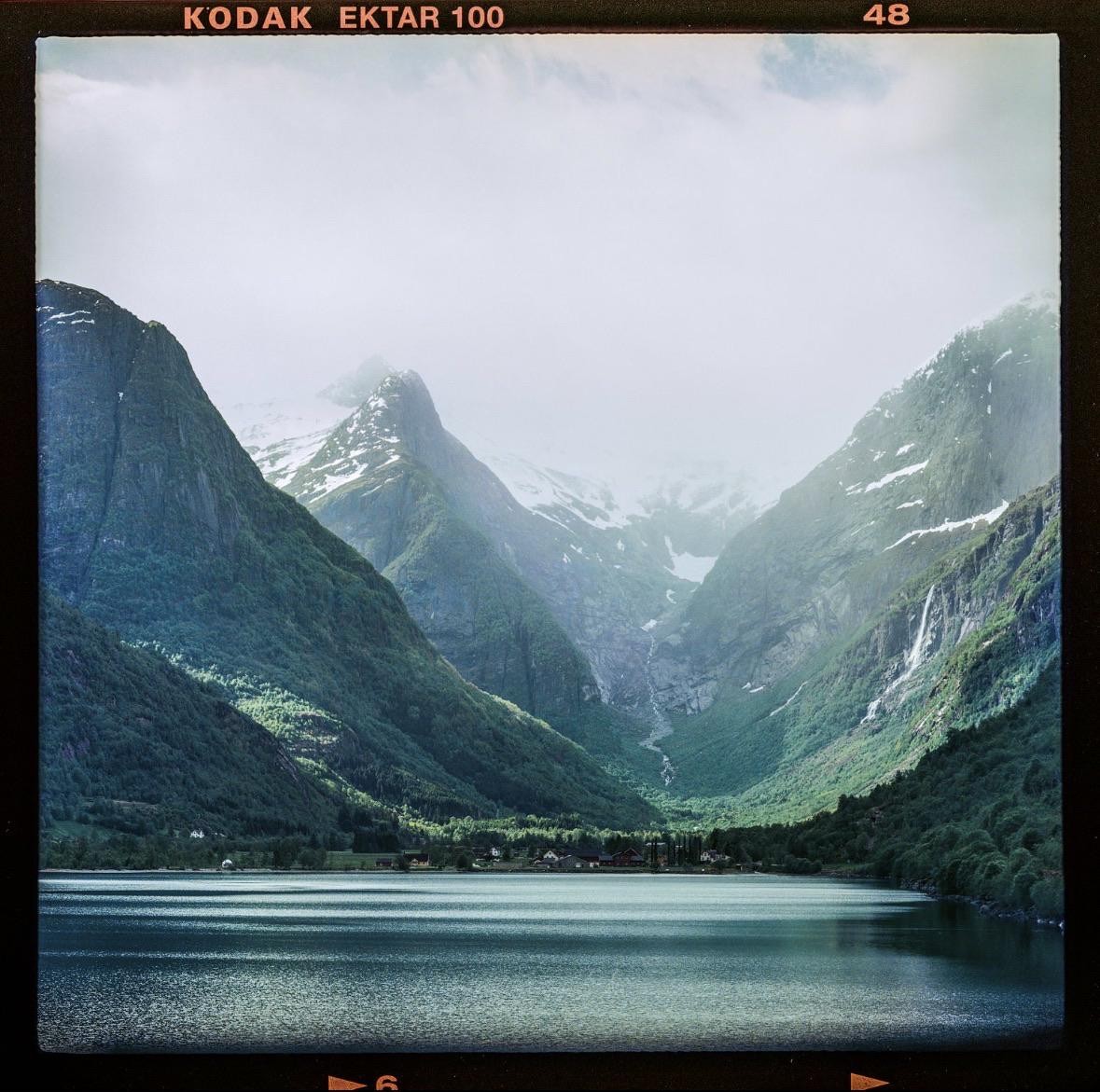 A body of water reflects the surrounding mountains, some of which have snow-capped peaks and waterfalls.