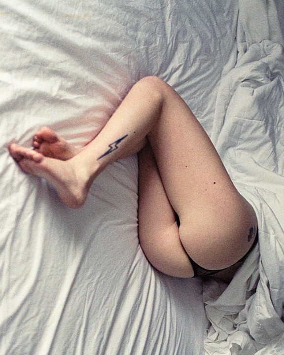 A woman's legs and buttocks are visible as she lies on white bedsheets, with a lightning bolt tattoo on her calf.