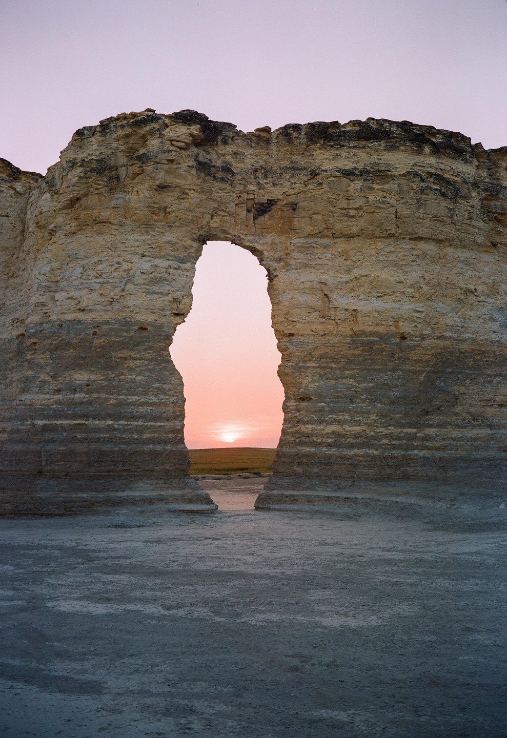 A sunset is visible through a natural rock archway in a flat landscape.