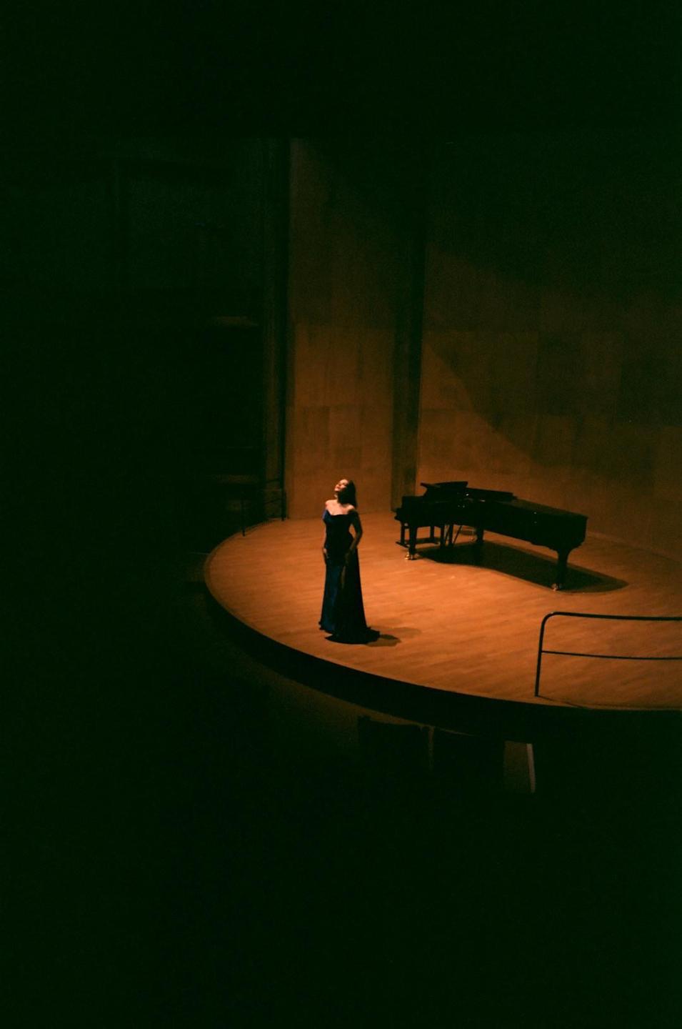 A woman in a long dress stands on a stage next to a grand piano, illuminated by a spotlight.