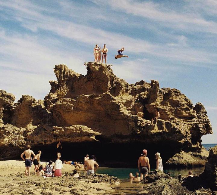 A man does a backflip off a large rock formation into the water while other people watch from the rocks and beach.