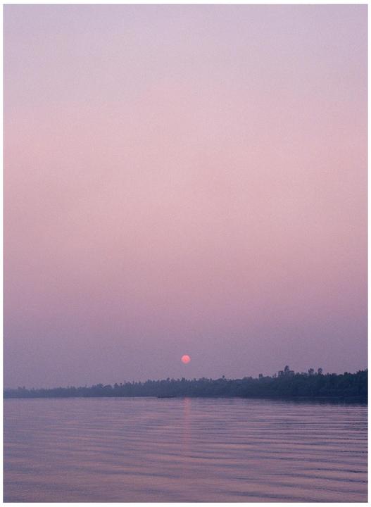 The sun sets over a body of water with a tree-lined shore.
