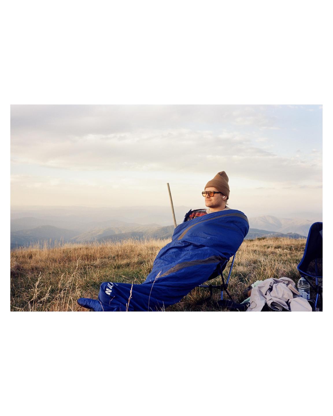 A man in a sleeping bag sits in a chair on a grassy mountain overlooking a valley.