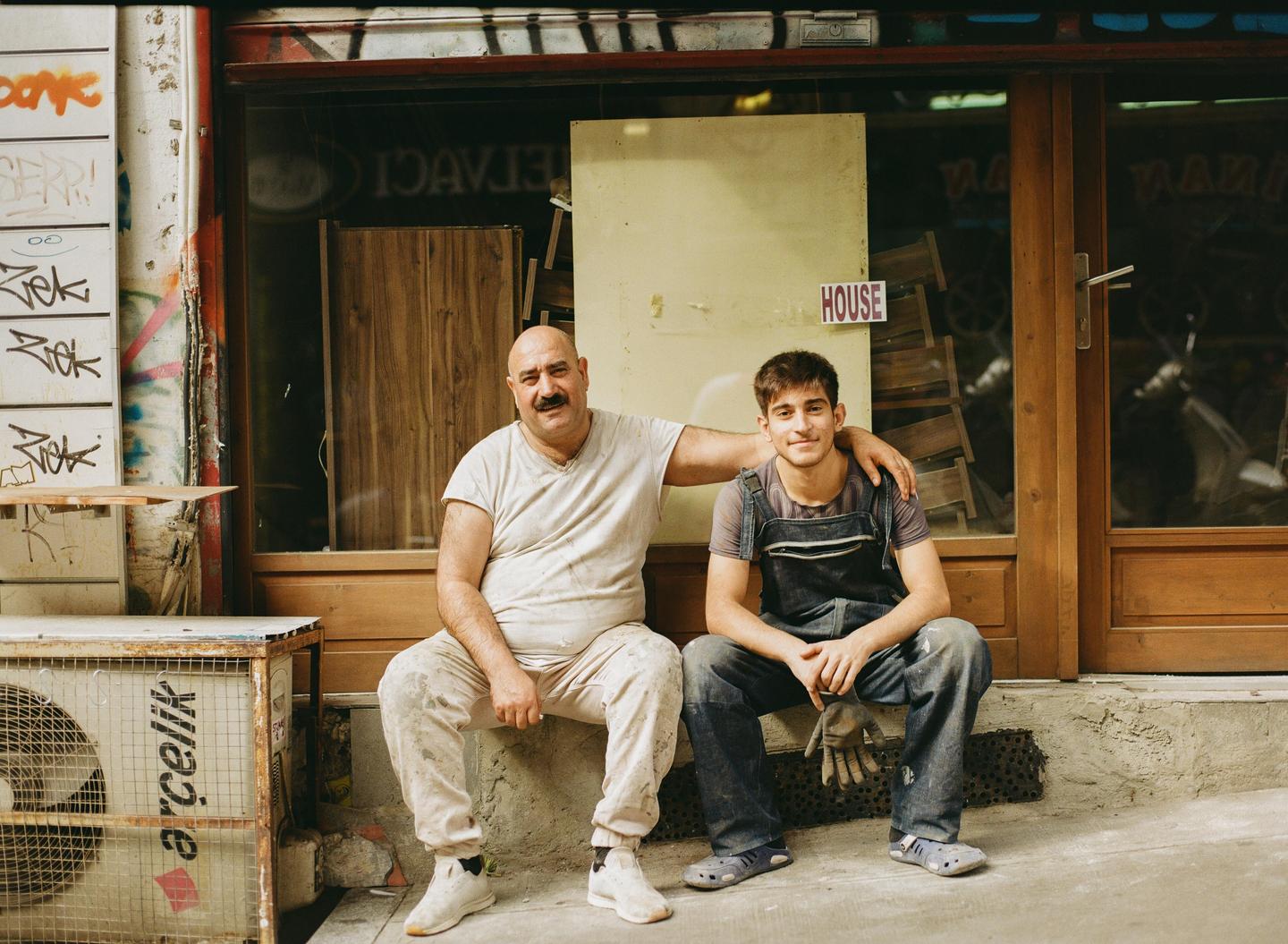 A man and a boy sit on a ledge in front of a storefront with graffiti.