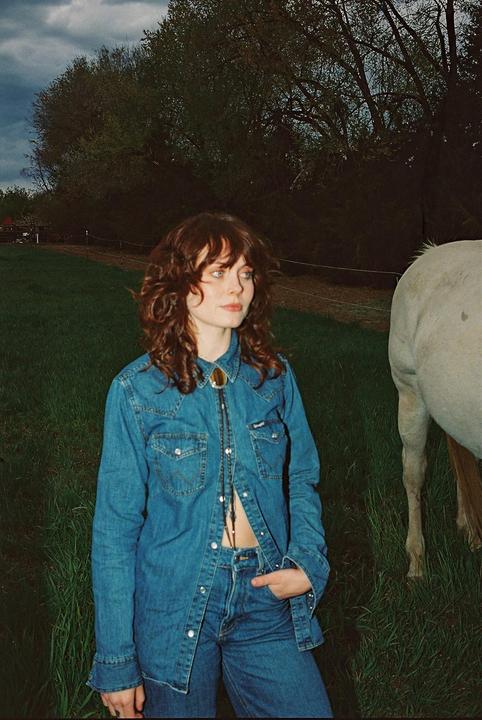 A woman in a denim shirt and jeans stands in a grassy field next to a horse.