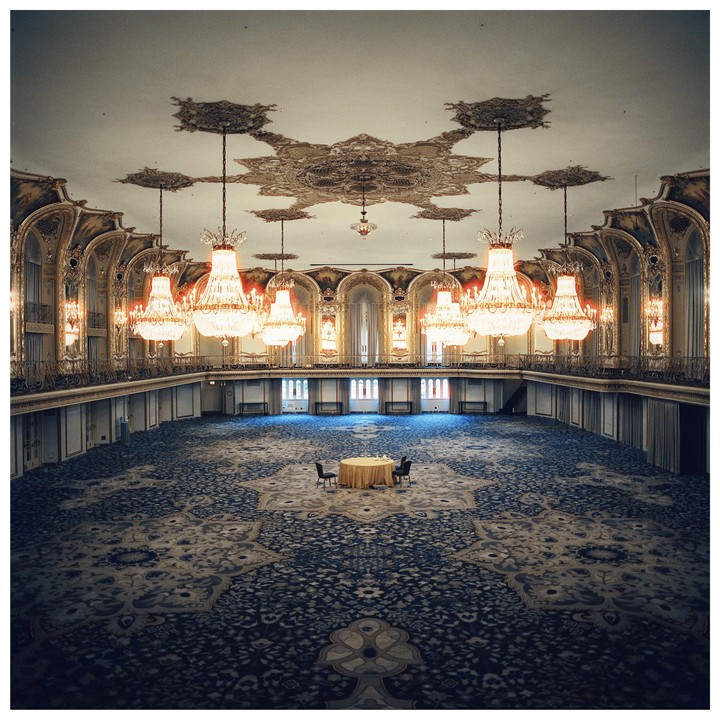 A grand ballroom with a high ceiling and ornate chandeliers features a single small table and two chairs in the center of a patterned carpet.