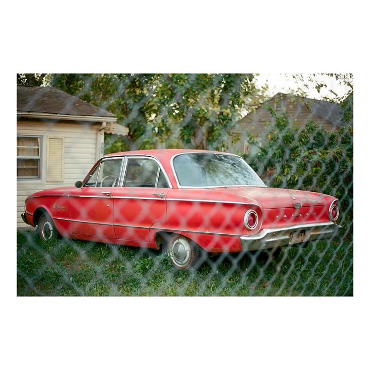 A red car is parked in a grassy yard behind a chain link fence.