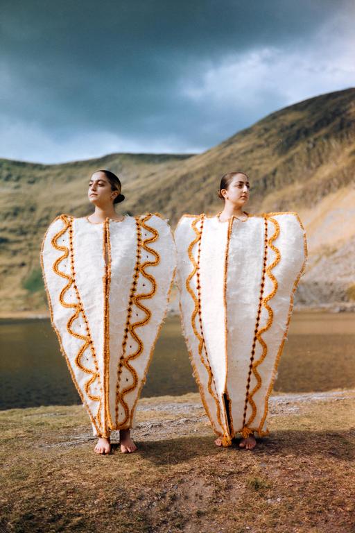Two women in elaborate white and gold costumes stand barefoot on a grassy bank beside a lake, with mountains and a cloudy sky in the background.