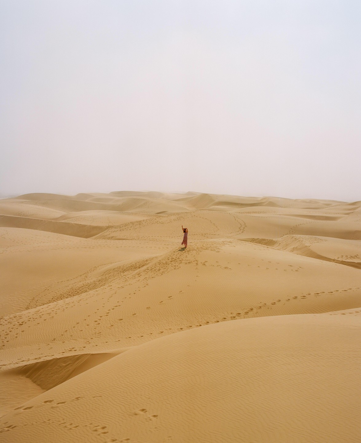 A woman in a pink dress stands on a sand dune with her arms raised, surrounded by many other sand dunes under a hazy sky.