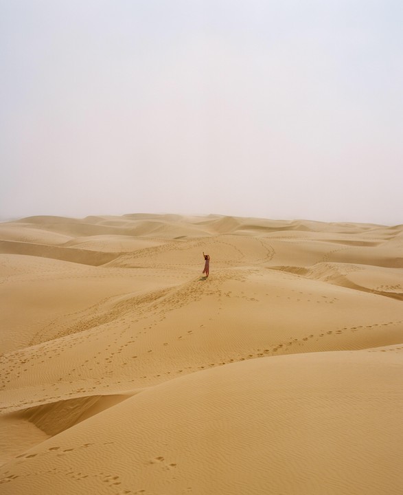 A woman in a pink dress stands on a sand dune with her arms raised, surrounded by many other sand dunes under a hazy sky.
