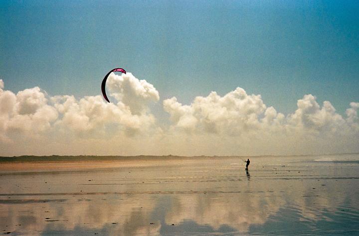 A person kitesurfs on a wide, wet beach under a cloudy sky.