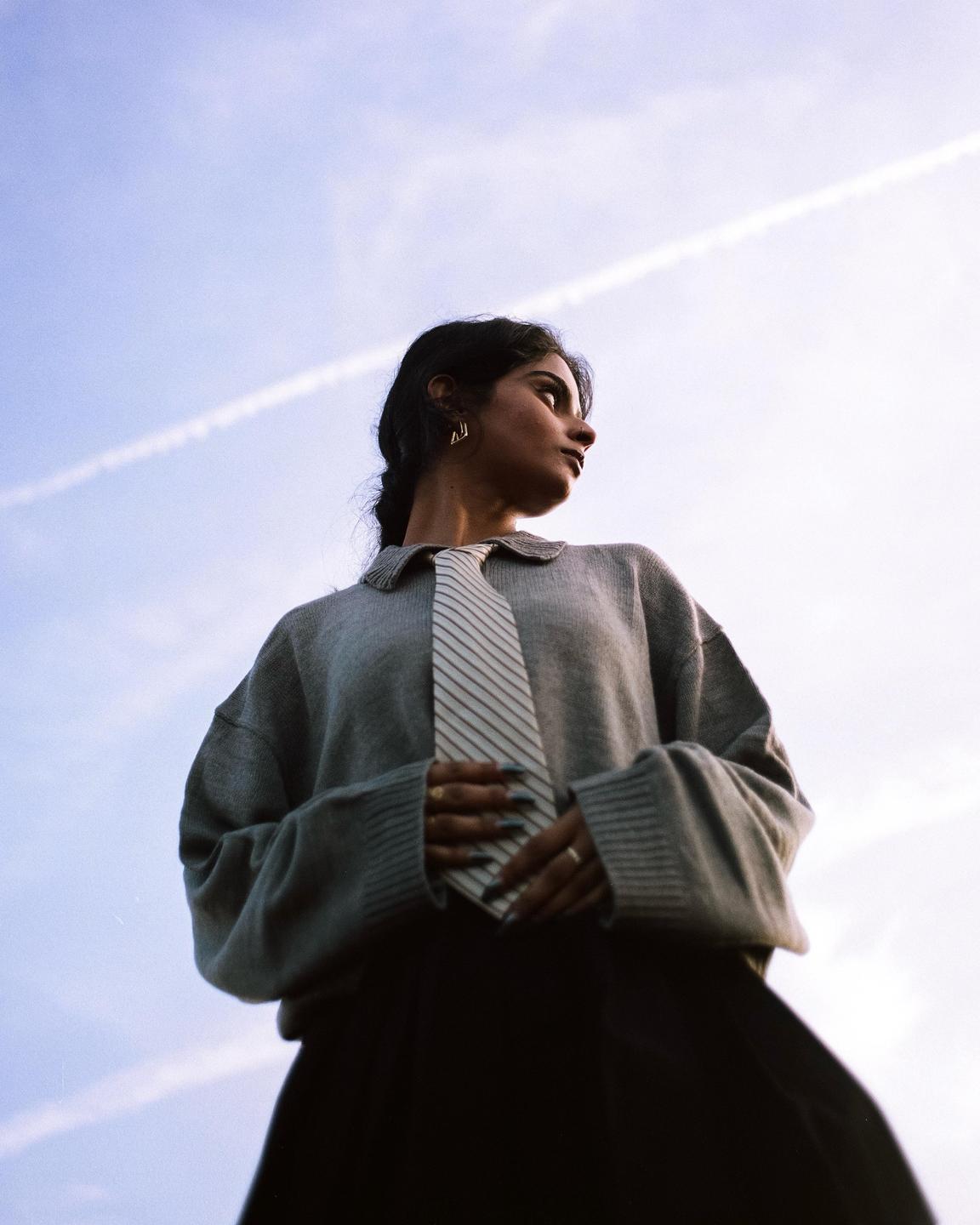 A woman wearing a sweater and a striped tie looks up at the sky, with a contrail visible in the background.