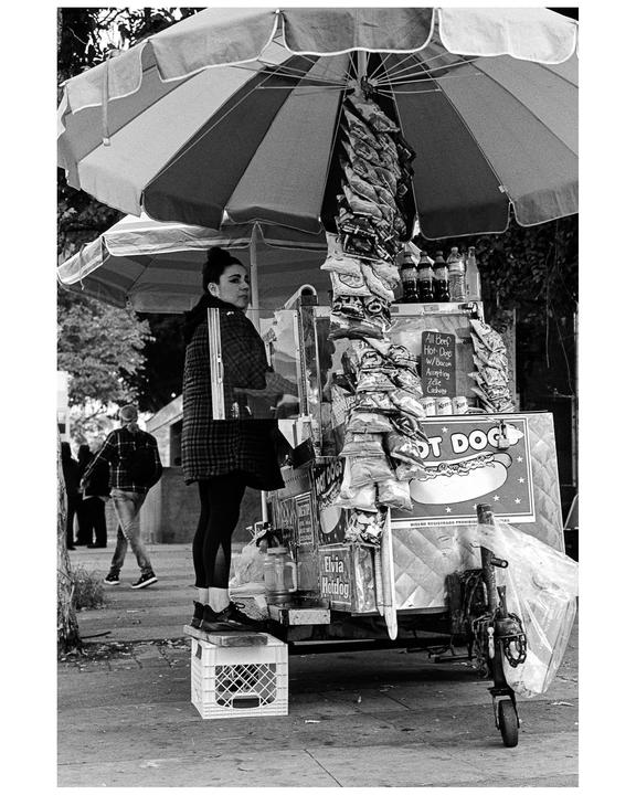 A woman stands on a crate behind a hot dog stand with an umbrella on a street.