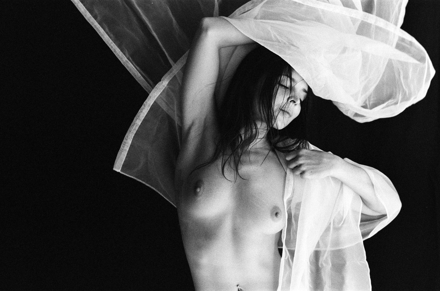 A topless woman with closed eyes is draped in sheer fabric against a dark background.