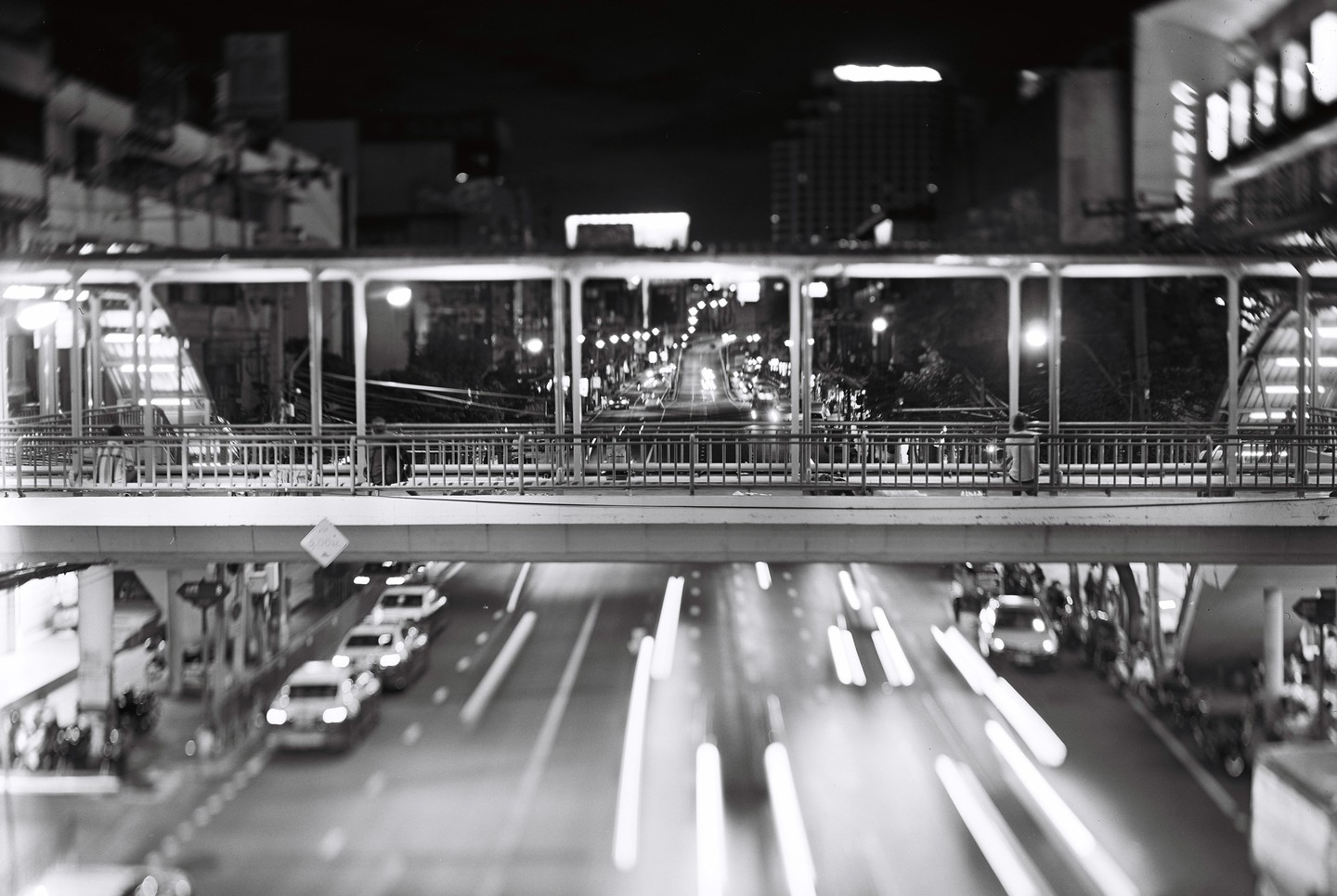 A black and white long exposure shows a pedestrian bridge over a busy street at night.