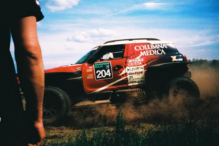 A red rally car kicks up dust on a dirt track under a blue sky, with a person's arm visible in the foreground.