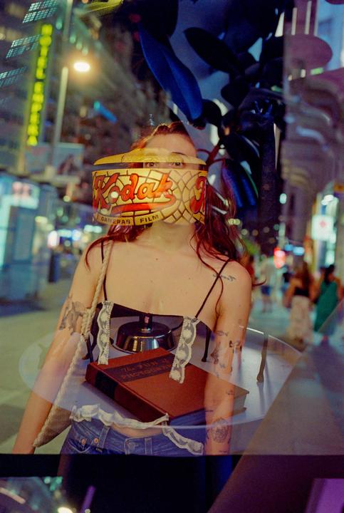 A woman with a Kodak film box on her face stands on a street at night.