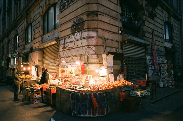 A street vendor sells fruit from a stall on a city corner at night, illuminated by several bright lights.
