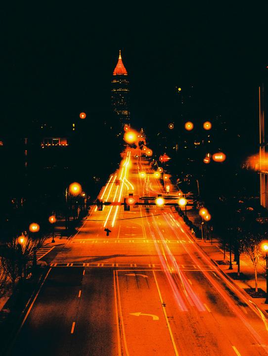 A long exposure shot of a city street at night shows car light trails leading towards a tall building with a pointed, illuminated red roof.