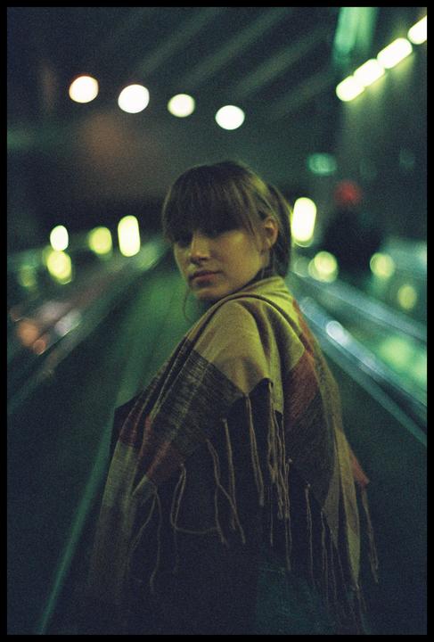 A woman wearing a patterned shawl looks over her shoulder while standing on an escalator at night.