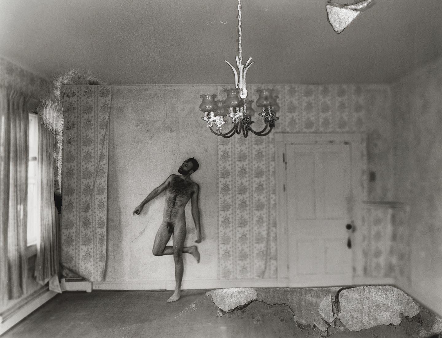 A nude man stands in a room with patterned wallpaper, a window, and a chandelier.