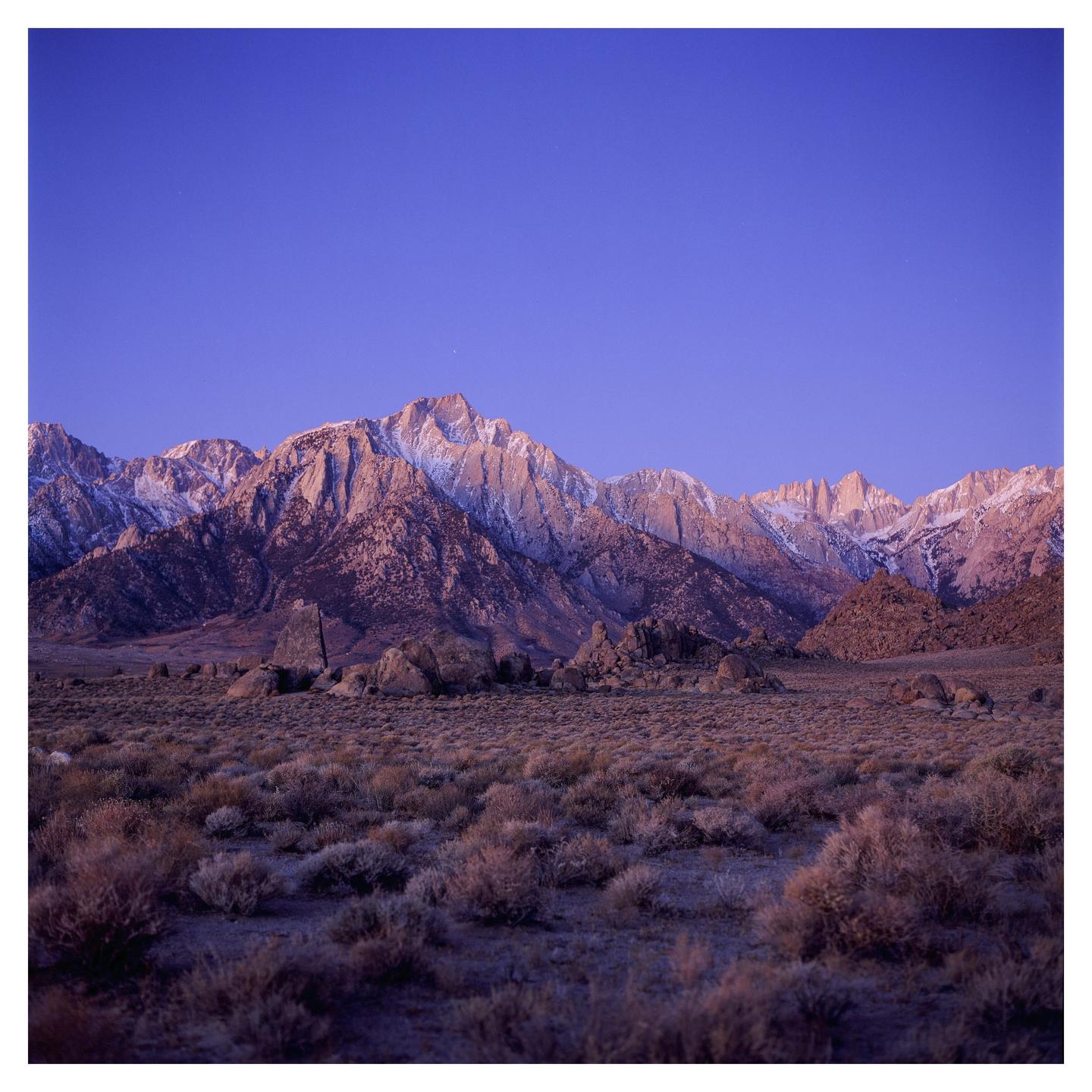 A vast desert landscape with sparse vegetation and large rocks leads to a snow-capped mountain range under a clear blue sky.