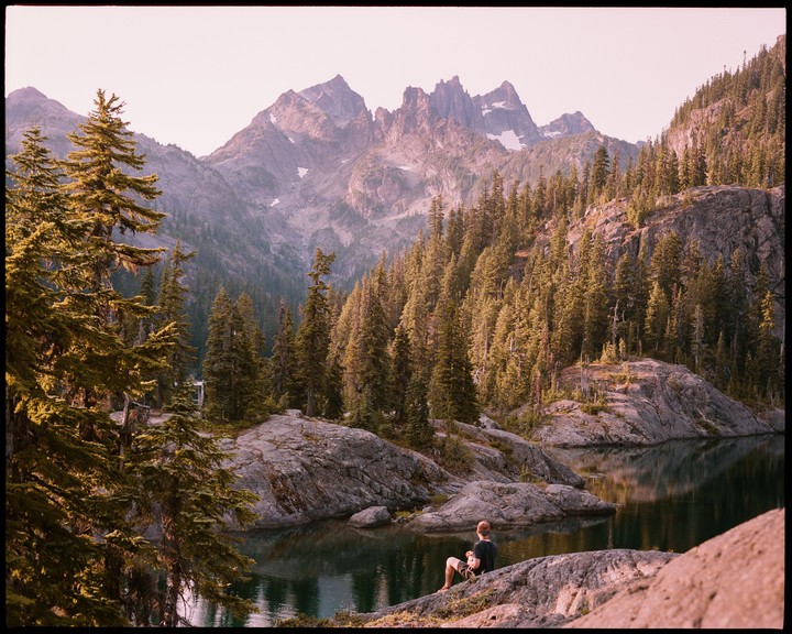 A man sits on a rock by a lake surrounded by pine trees and mountains.