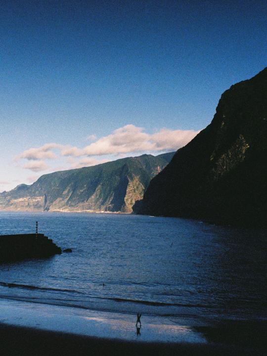 A person stands on a beach with mountains and a body of water in the background.