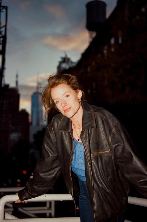 A woman in a leather jacket stands on a balcony with a city skyline in the background at dusk.