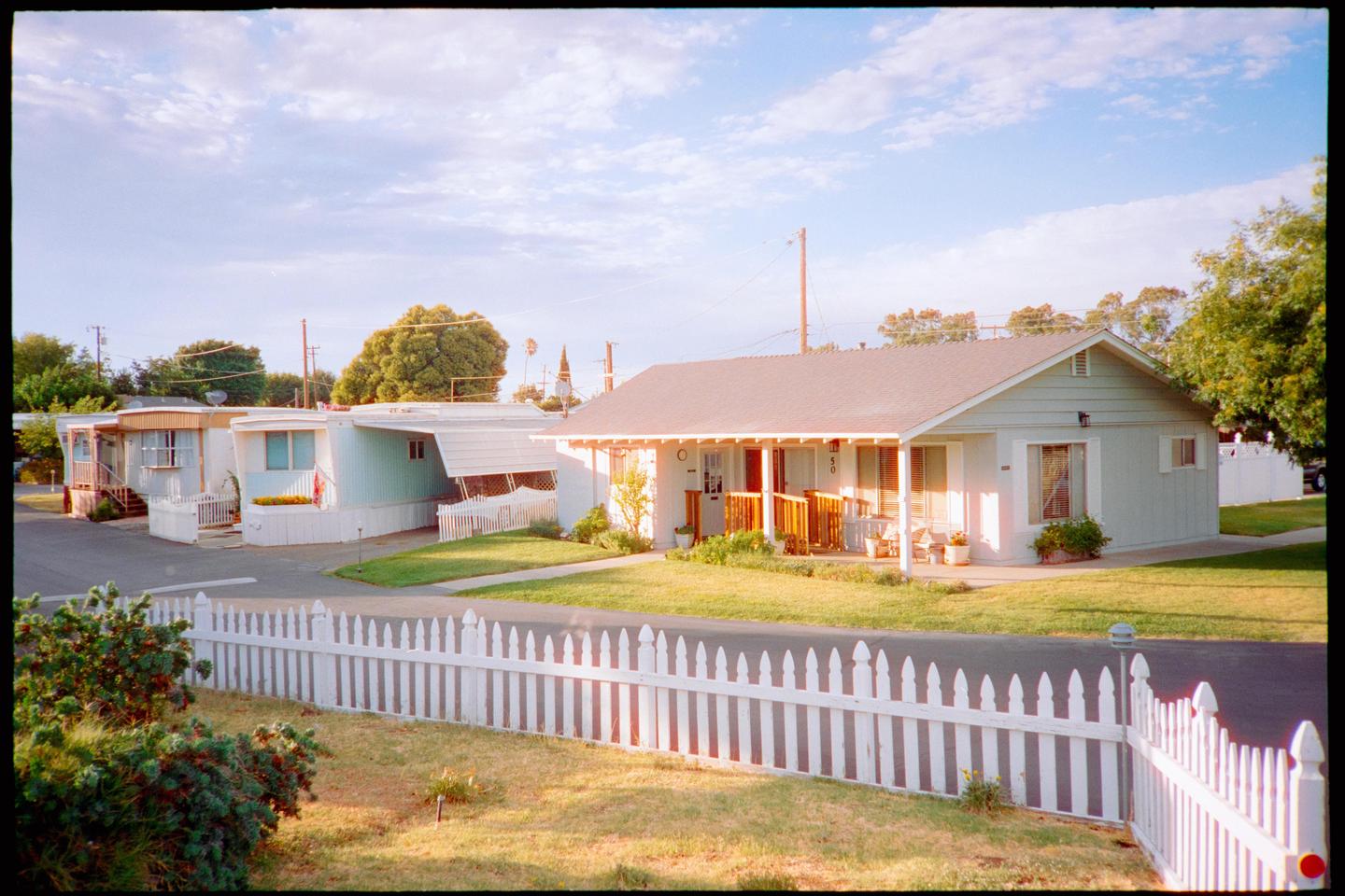 A white picket fence runs along a paved road with houses and trees under a partly cloudy sky.