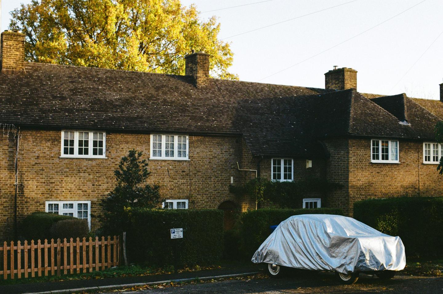 A car covered with a silver tarp is parked in front of a brick house with a hedge and a wooden fence.