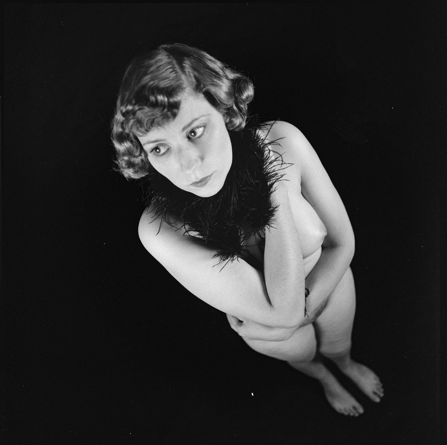 A woman with curled hair and a feather boa poses nude against a dark background.