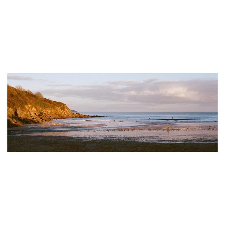 A person walks on a wide beach with a cliff on the left and the ocean on the right under a cloudy sky.