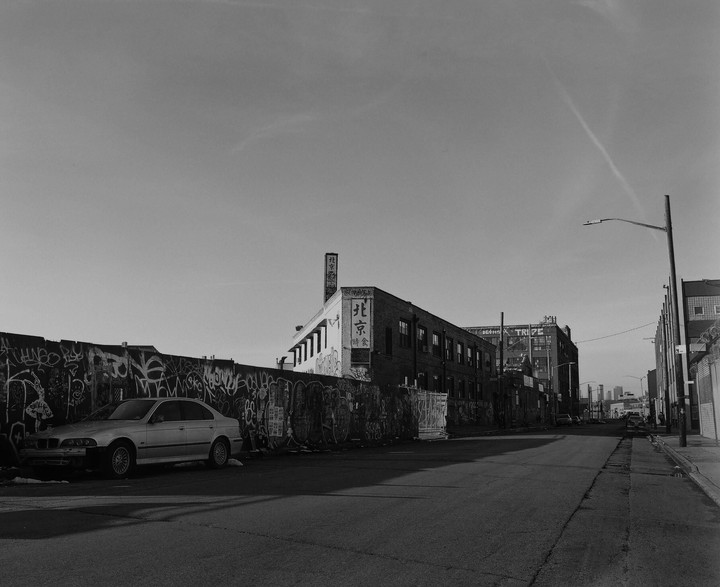 A street with a graffiti-covered wall and a parked car leads to a building with Chinese characters on its facade.