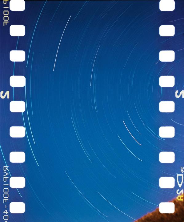 A blue night sky with star trails is visible through the sprocket holes of a film strip.