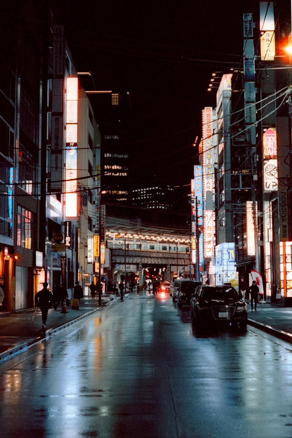 A street in Tokyo is wet from rain at night, with neon signs illuminating the buildings and a train passing on an elevated track.