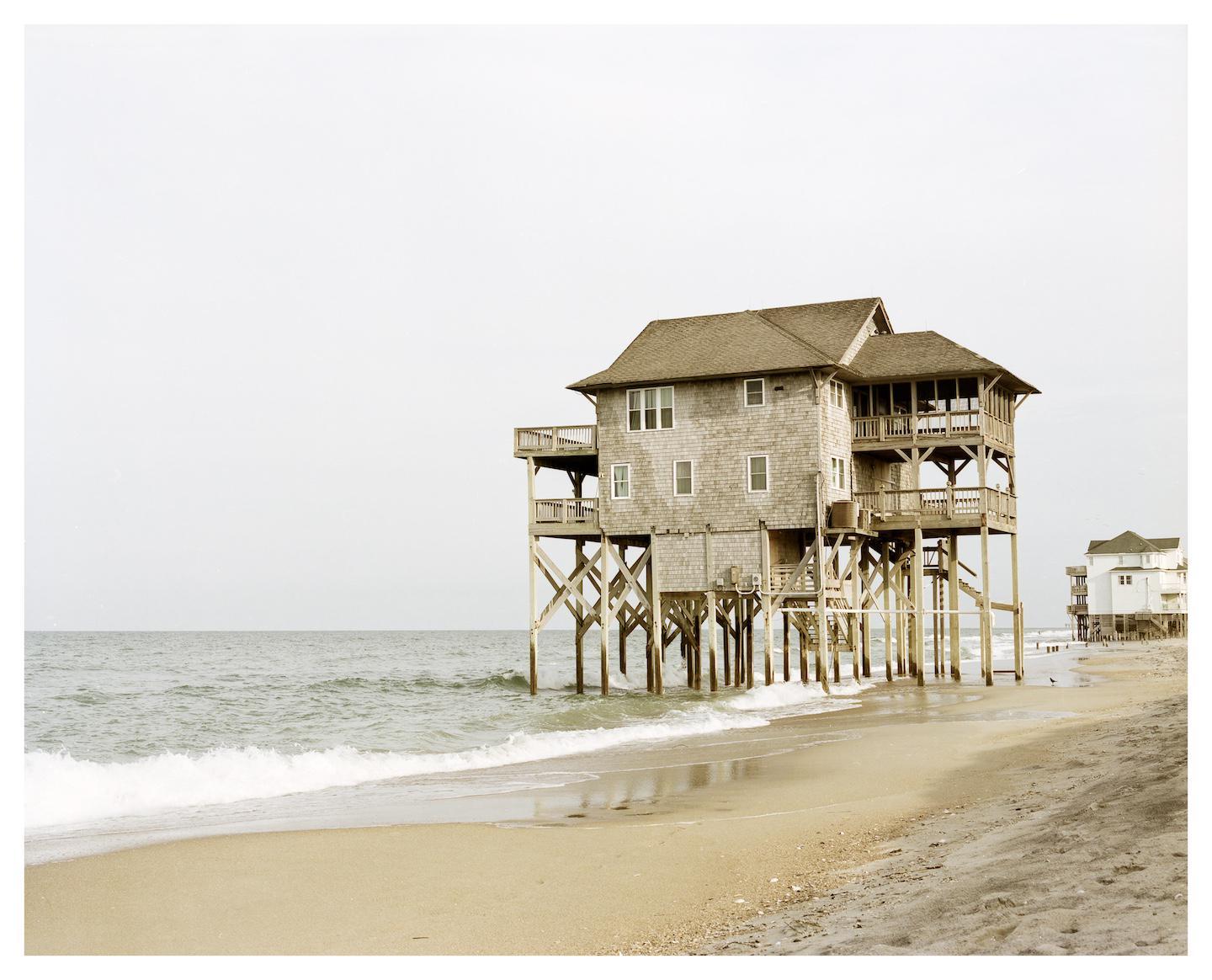 A large house on stilts stands on a sandy beach next to the ocean under a bright sky.
