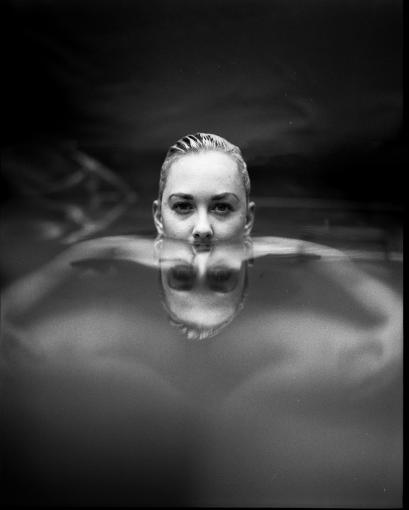A woman's face is visible above the water, with her reflection appearing below the surface.