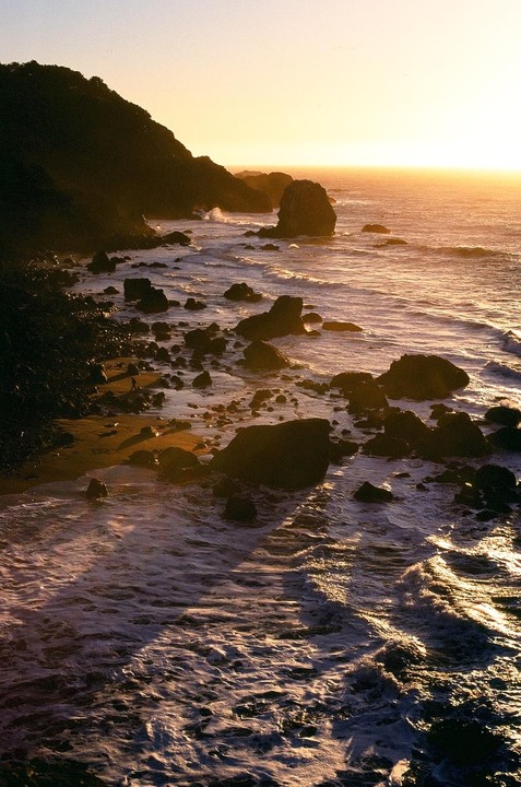 The sun sets over a rocky coastline with waves crashing against the shore.